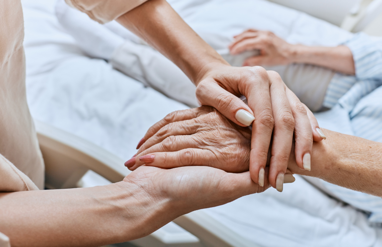 End of Life Care Two hands clasped together, one aged and the other younger, on a hospital bed.