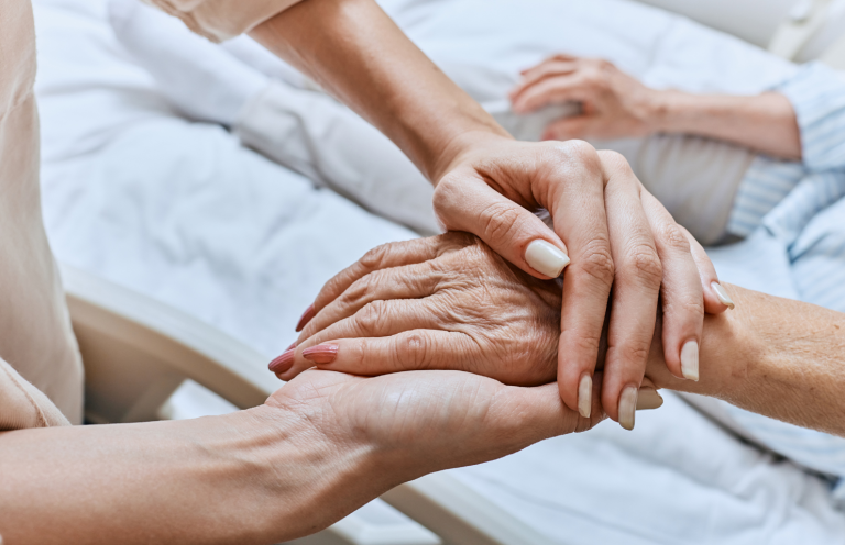Recognising a Deteriorating Patient A caregiver gently holds the hand of an elderly person in a bed.