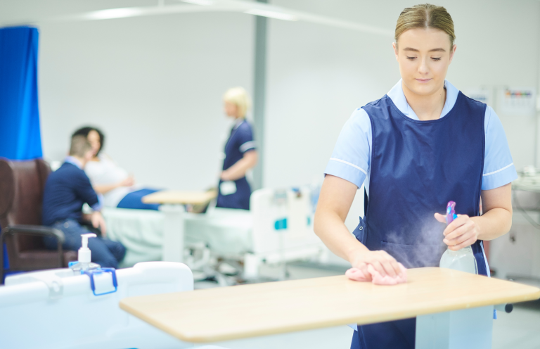 A healthcare worker cleans a table in a hospital setting while patients are treated nearby.