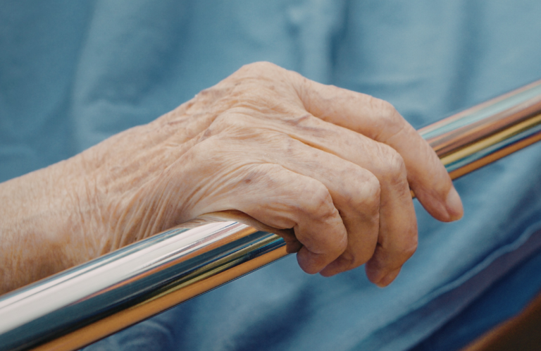 An elderly hand resting on a metal railing, with light blue fabric in the background.