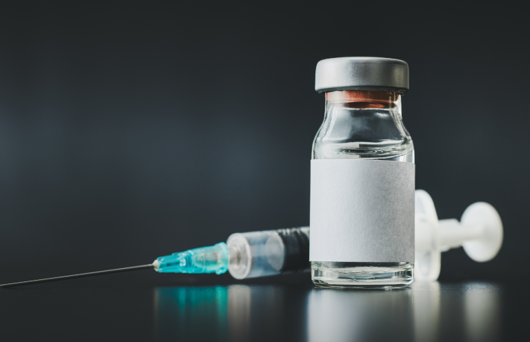 A vial of medicine beside a syringe and needle, set against a dark background.