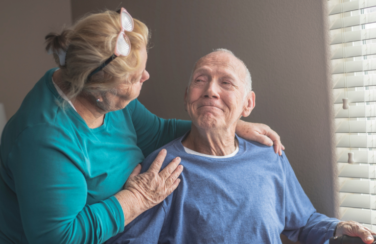 Dementia Awareness Training A woman tenderly smiles at an elderly man seated indoors by a window.