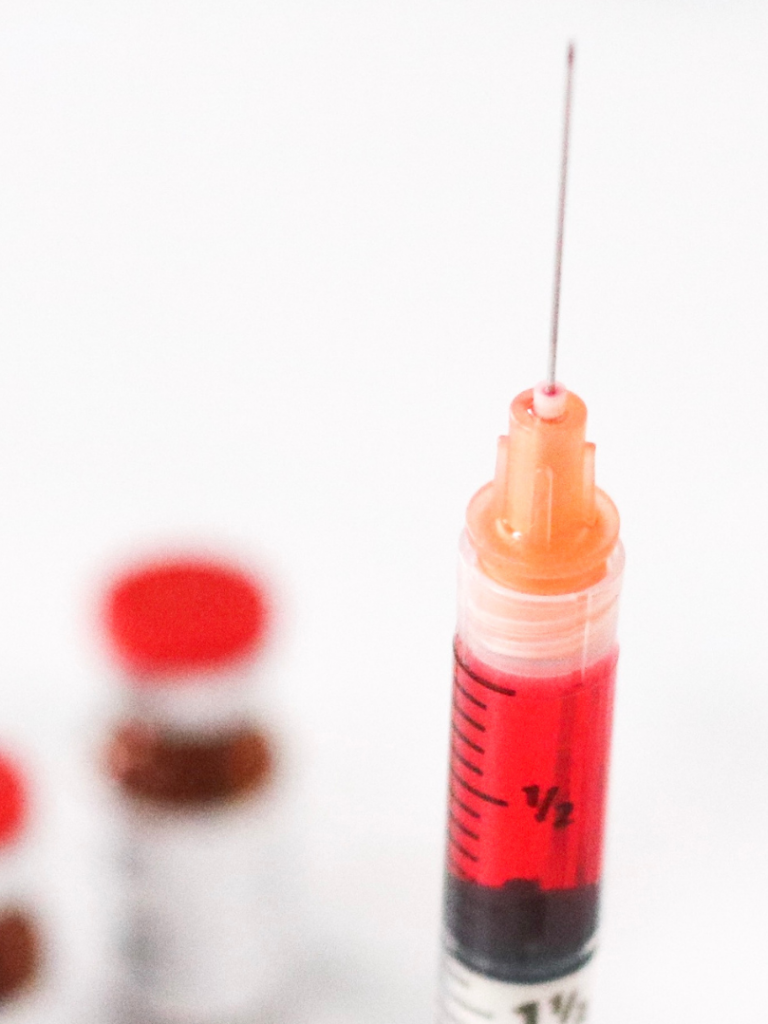 Syringe filled with red liquid, with vials in the background.
