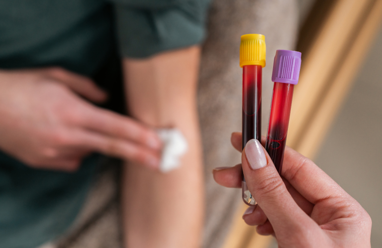 A hand holds two blood sample tubes next to a person’s arm being cleaned.