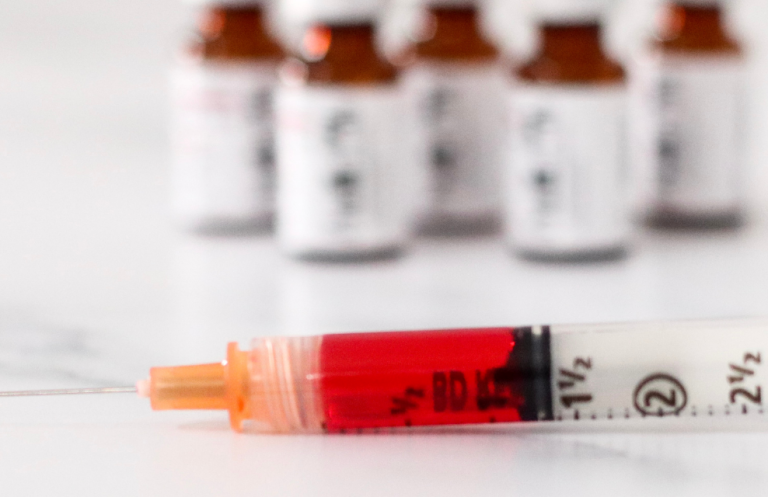 B12 and Injection Techniques Syringe filled with red liquid in foreground, with brown vials blurred in the background.